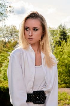 Young blonde woman in white skirt and shirt in the old sunny summer city park. Fashion woman. Young woman's modern portrait.