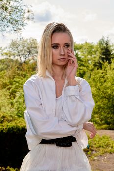 Young blonde woman in white skirt and shirt in the old sunny summer city park. Fashion woman. Young woman's modern portrait.