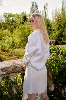 Young blonde woman in white skirt and shirt in the old sunny summer city park. Fashion woman. Young woman's modern portrait.