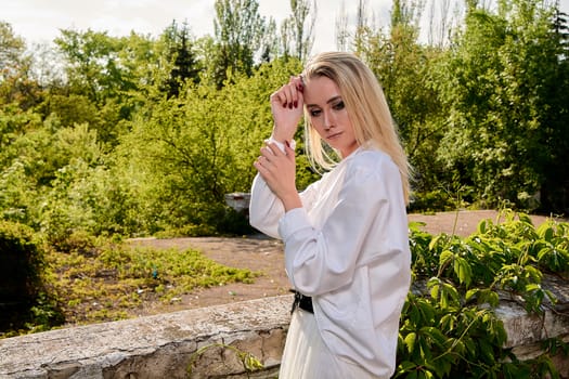 Young blonde woman in white skirt and shirt in the old sunny summer city park. Fashion woman. Young woman's modern portrait.