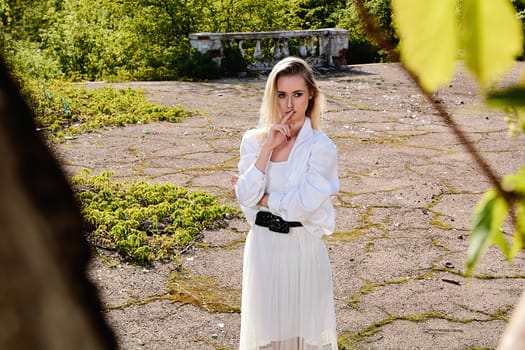 Young blonde woman in white skirt and shirt in the old sunny summer city park. Fashion woman. Young woman's modern portrait.