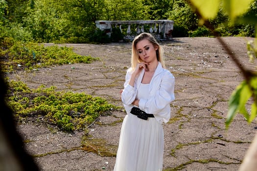 Young blonde woman in white skirt and shirt in the old sunny summer city park. Fashion woman. Young woman's modern portrait.