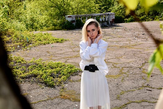 Young blonde woman in white skirt and shirt in the old sunny summer city park. Fashion woman. Young woman's modern portrait.