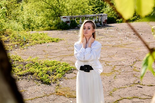 Young blonde woman in white skirt and shirt in the old sunny summer city park. Fashion woman. Young woman's modern portrait.