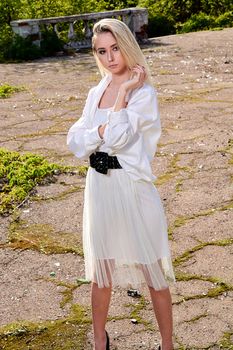 Young blonde woman in white skirt and shirt in the old sunny summer city park. Fashion woman. Young woman's modern portrait.