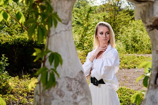 Young blonde woman in white skirt and shirt in the old sunny summer city park. Fashion woman. Young woman's modern portrait.