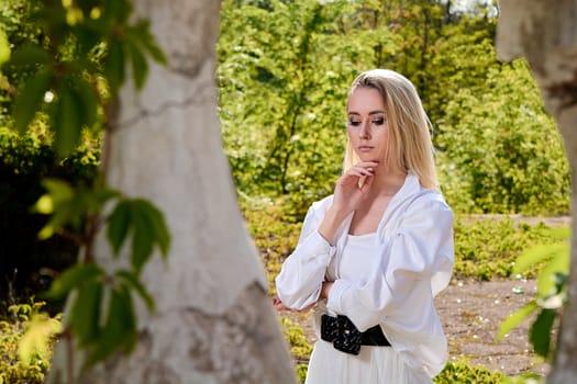 Young blonde woman in white skirt and shirt in the old sunny summer city park. Fashion woman. Young woman's modern portrait.