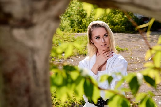 Young blonde woman in white skirt and shirt in the old sunny summer city park. Fashion woman. Young woman's modern portrait.