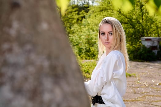 Young blonde woman in white skirt and shirt in the old sunny summer city park. Fashion woman. Young woman's modern portrait.