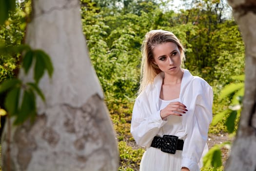 Young blonde woman in white skirt and shirt in the old sunny summer city park. Fashion woman. Young woman's modern portrait.