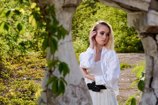 Young blonde woman in white skirt and shirt in the old sunny summer city park. Fashion woman. Young woman's modern portrait.