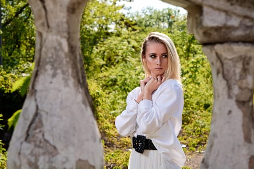 Young blonde woman in white skirt and shirt in the old sunny summer city park. Fashion woman. Young woman's modern portrait.