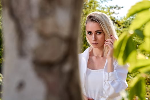 Young blonde woman in white skirt and shirt in the old sunny summer city park. Fashion woman. Young woman's modern portrait.