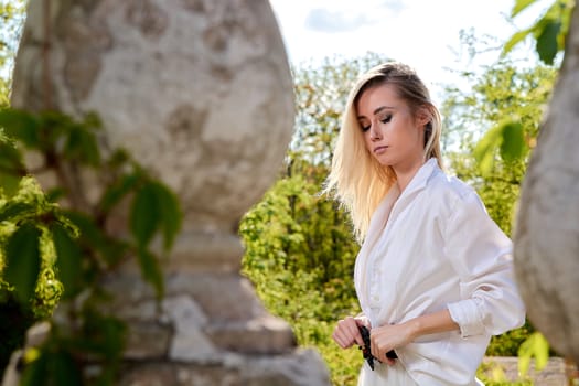 Young blonde woman in white skirt and shirt in the old sunny summer city park. Fashion woman. Young woman's modern portrait.