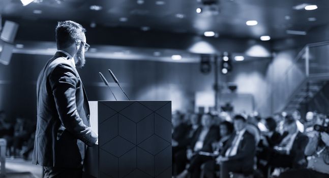 Speaker giving a talk on corporate business conference. Unrecognizable people in audience at conference hall. Business and Entrepreneurship event. Blue toned grayscale image.