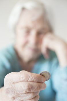 Sad elderly woman sitting at the table at home and looking miserably at only remaining coin from pension in her hand. Unsustainability of social transfers and pension system. Focus on coin.