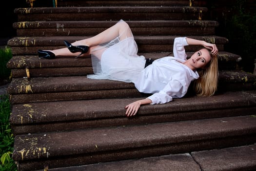 Young blonde woman in white skirt and shirt sits on the steps of the old granite stair. Fashion woman. Young woman modern portrait.