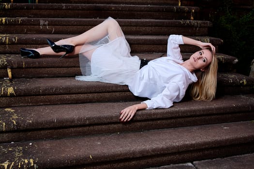 Young blonde woman in white skirt and shirt sits on the steps of the old granite stair. Fashion woman. Young woman modern portrait.