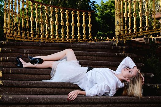 Young blonde woman in white skirt and shirt sits on the steps of the old granite stair. Fashion woman. Young woman modern portrait.