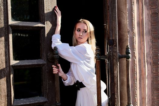 Young blonde woman in white skirt and shirt stands in the old wooden doorway. The old wooden door. Fashion woman. Young woman modern portrait.