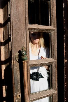 Young blonde woman in white skirt and shirt stands in the old wooden doorway. The old wooden door. Fashion woman. Young woman modern portrait.