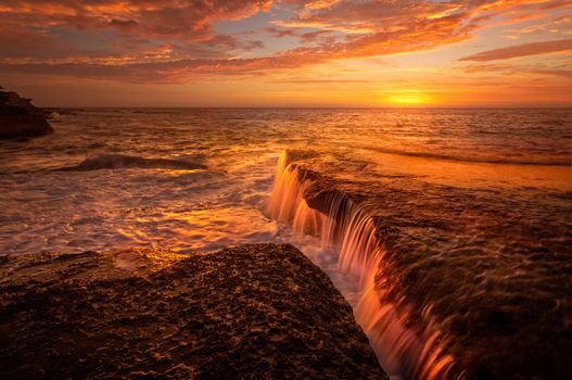 Tidal waters flow off rocks creating glowing orange waterfalls in the summer sunrise on the coast near Sydney Australia