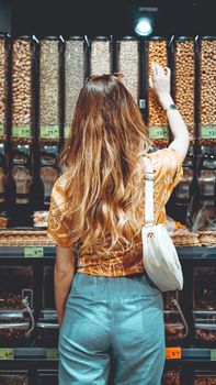 Young positive woman standing next to containers with nuts and picking selected in organic store