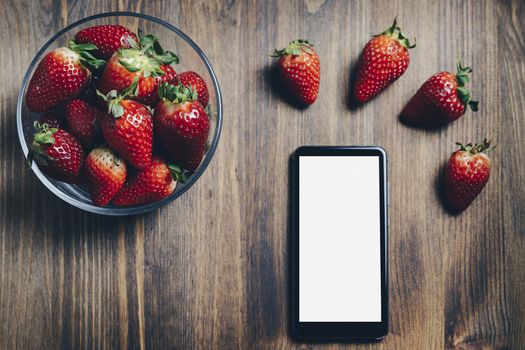Juicy fresh strawberries in a glass bowl and a phone on old wooden background, healthy sweet food, vitamins and fruity concept. Top view, copy space for text