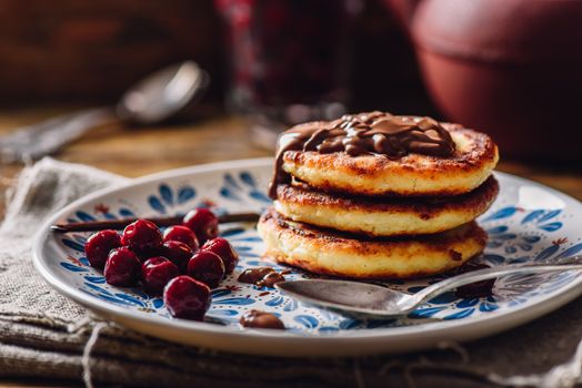 Stack of Quark Pancakes with Hazelnut Paste, Frozen Cherry and Vanilla Pod. Tea Pot with Spoons and Glass of Berries on Background.