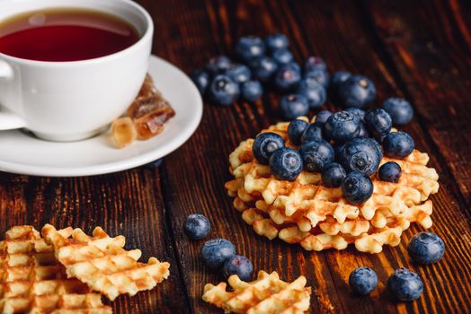 Cup of Tea with Blueberries on the Top of the Waffles Stack and Other Scattered on Wooden Background.