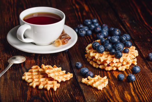 Cup of Tea with Blueberries on the Top of the Belgian Waffles Stack and Other Scattered on Wooden Background.