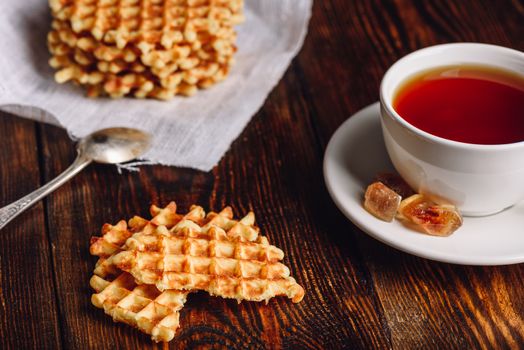 White Cup of Tea with Waffles Stack on Napkin and Pieces of Waffle on Wooden Surface and Spoon.