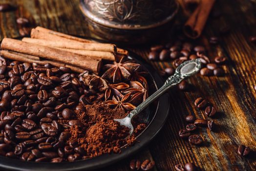 Coffee Beans with Spooonful of Ground Coffee, Cinnamon Sticks and Chinese Star Anise on Metal Plate. Some Beans Scattered on Wooden Table and Cezve on Backdrop.