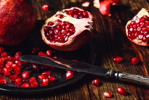 Opened Pomegranate and Whole One wiith Seeds on Metal Plate and Vintage Knife. Some Seeds and Pieces Scattered on Wooden Background.