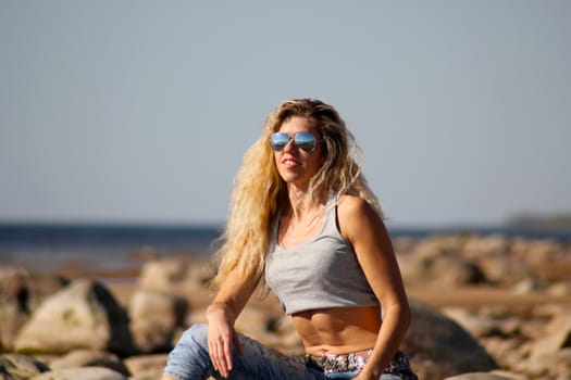 Tanned blonde girl in sunglasses and jeans sits on the rocks on the beach on a sunny day.