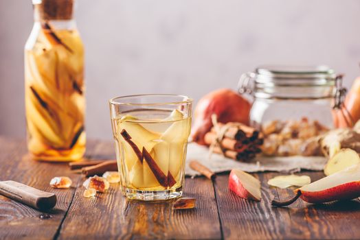 Glass of Water Flavored with Sliced Pear, Cinnamon Stick, Ginger Root and Some Sugar. Ingredients on Wooden Table and Bottle of beverage on Backdrop.