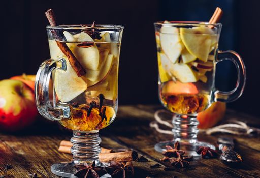 Two Glasses of Christmas Spiced Beverage with Sliced Apple, Clove, Cinnamon, Anise Star and Dark Candy Sugar. All Ingredients and Some Kitchen utensils on Wooden Table.Vertical.