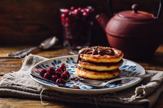 Russian Quark Pancakes with Chocolate Topping and Frozen Cherry. Tea Pot with Spoons and Glass of Berries on Background.