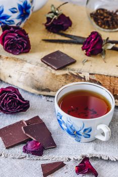 Cup of Black Tea with Chocolate Bars, Dry Rosebuds and Teapot with Jar and Rusty Scissors on Board. Vertical Orientation.