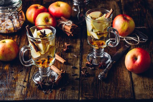 Glasses of Apple Spiced Drink with Clove, Cinnamon, Anise Star and Dark Candy Sugar. All Ingredients and Some Kitchen utensils on Wooden Table. Vertical.