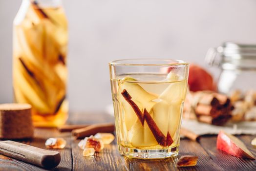 Glass of Detox Water with Sliced Pear, Cinnamon Stick, Ginger Root and Some Sugar. Ingredients on Wooden Table and Bottle of beverage on Backdrop.