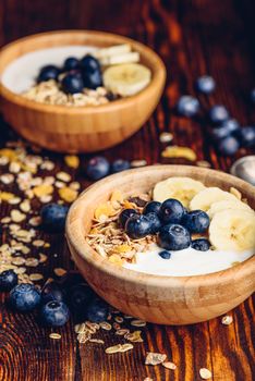 Two Bowl of Granola, Banana, Blueberry and Greek Yoghurt. Scattered Ingredients on Wooden Table. Vertical Orientation.