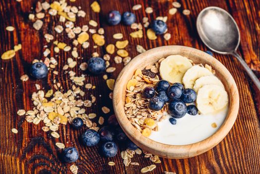 Bowl of Granola, Banana, Blueberry and Greek Yoghurt. Scattered Ingredients and Spoon on Wooden Table.