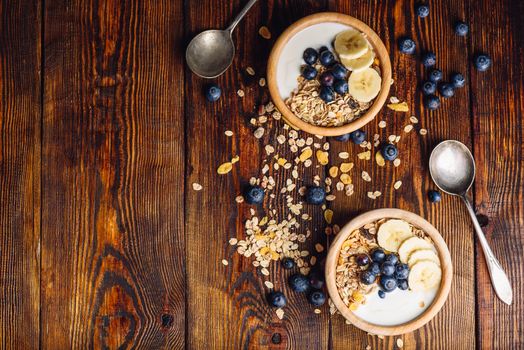 Two Bowl of Granola, Banana, Blueberry and Greek Yoghurt. Scattered Ingredients on Wooden Table. View from Above and Copy Space.