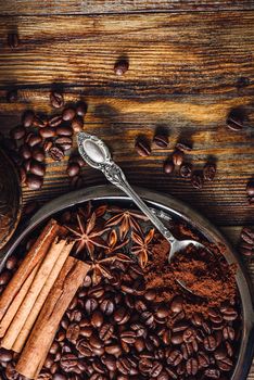 Coffee Beans with Spooonful of Ground Coffee, Cinnamon Sticks and Star Anise on Plate. Some Beans Scattered on Wooden Table. View from Above. Vertical Orientation.