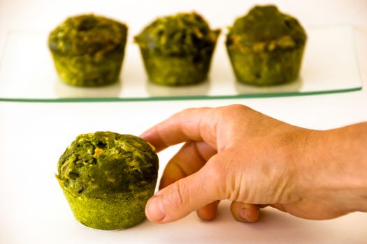 Three green muffins on a glass tray at the background and a hand catching another one at the foreground