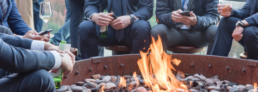 Panorama view group of Caucasian businessmen in formal dress gathering over the round fire pit after work at upscale bar in Chicago. Business people watches, leather shoes drinking beer, wine