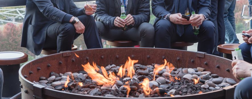 Panorama view group of Caucasian businessmen in formal dress gathering over the round fire pit after work at rooftop bar in Chicago. Business people watches, leather shoes drinking beer, wine