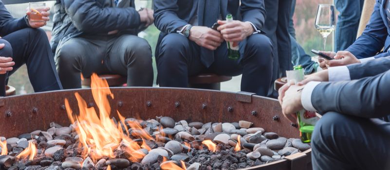 Panorama view group of Caucasian businessmen in formal dress gathering over the round fire pit after work at upscale bar in Chicago. Business people watches, leather shoes drinking beer, wine