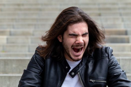 Portrait of a handsome bearded man with long brown hair, dressed in trendy clothes, sitting on the steps and yawning, mouth wide open, with a copy space for advertising content.